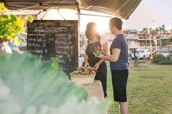 Two people standing in front of a market stall of vegetables