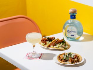 Don Julio tequila bottle next to a margarita and tacos with yellow wall and orange chair in the background. (Image: Nikki To)