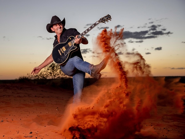 A man with a cowboy hat and a guitar kicking red sand with his boots.