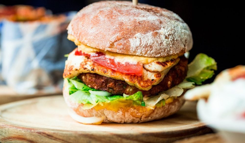 Close up image of a freshly flame grilled vegetarian halloumi cheeseburger on a wooden counter at an outdoor food market. This burger is loaded with a vegetarian burger meat substitute, fresh salad, melted halloumi cheese and spanish onion and tomato. The burger is sandwiched between glazed buns.