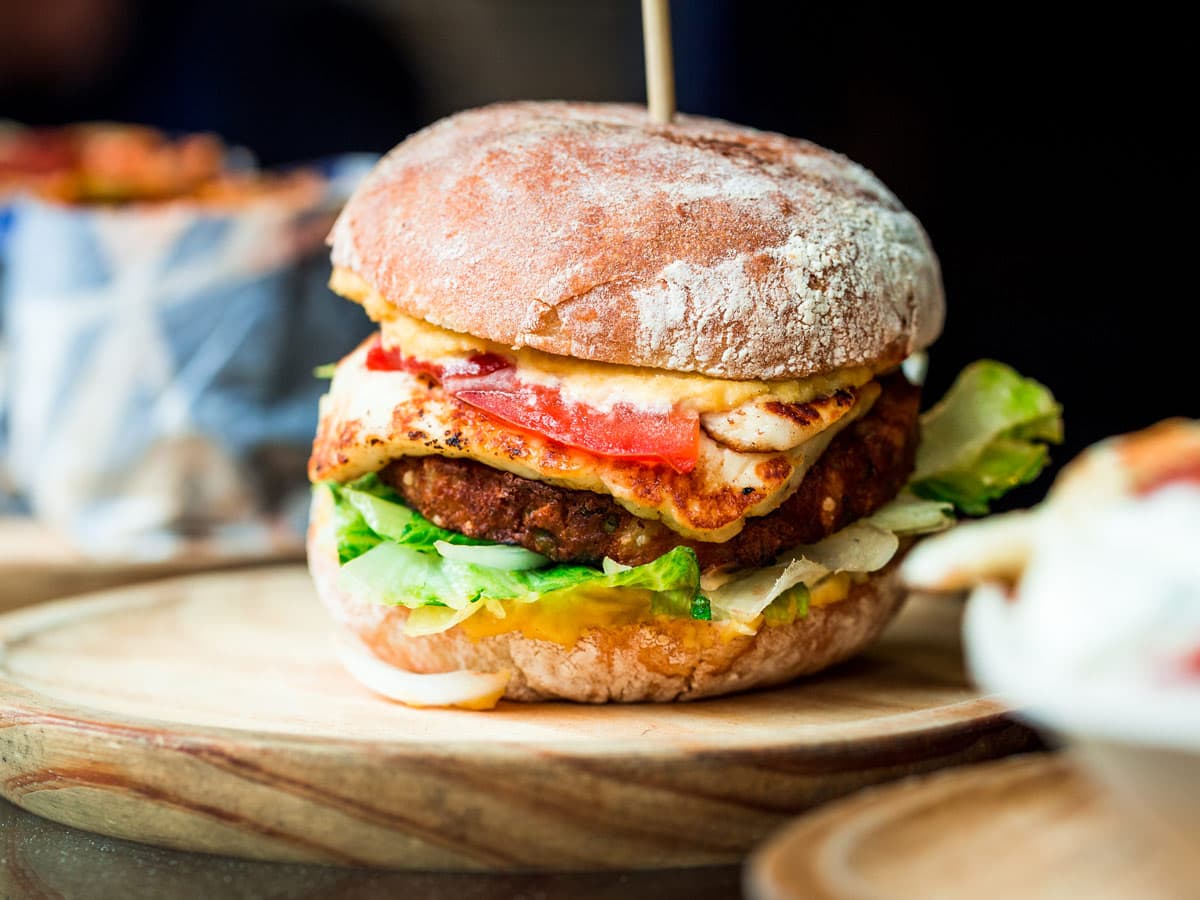 Close up image of a freshly flame grilled vegetarian halloumi cheeseburger on a wooden counter at an outdoor food market. This burger is loaded with a vegetarian burger meat substitute, fresh salad, melted halloumi cheese and spanish onion and tomato. The burger is sandwiched between glazed buns.