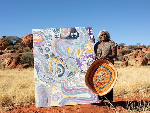Puna Yanima with one of her baskets for Tjanpi Desert Weavers and her painting Antara (2022). Photo by Tim Coad, courtesy the artist and Mimili Maku Arts, DAAF 2022