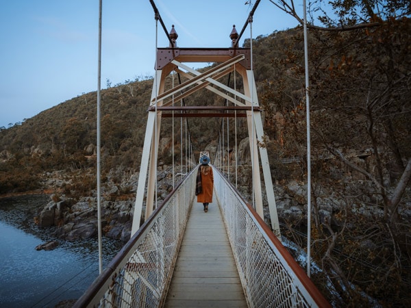 A girl with an orange coat and blue hat walking on a suspension bridge above the water.