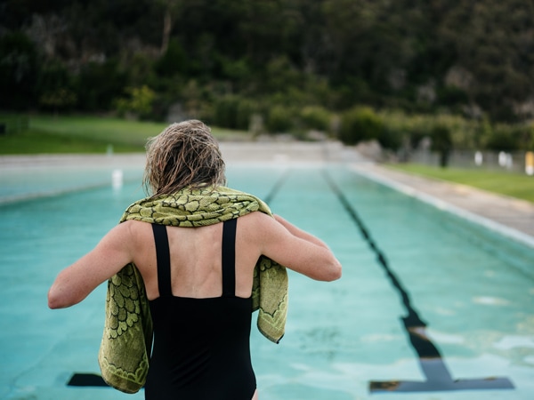 A blonde woman with black bathers and a green towel over her shoulders standing in front of a swimming pool.