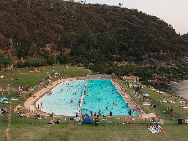 A blue swimming pool in the middle of the Cataract Gorde reserve with people bathing, tanning, and resting.