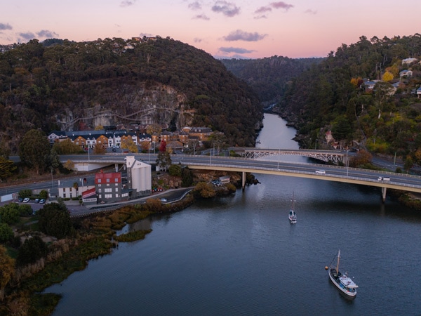 A bridge over a river with boats surrounded by a town and mountains at sunset.