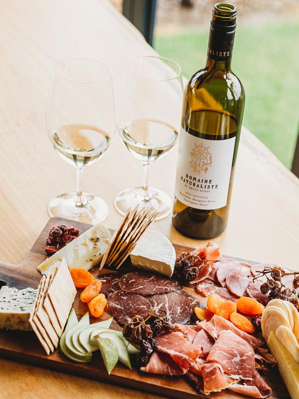 Two glasses of white wine with a bottle of wine and a charcuterie and cheese platter with dried fruit on a wooden table.