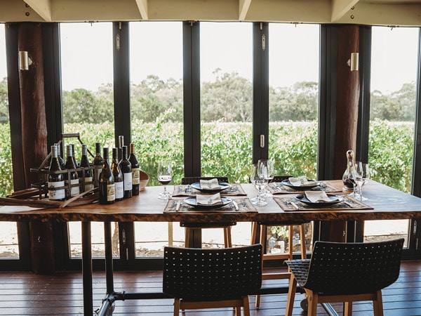 A table with chairs, plates, and wine bottles indoors in front of a vineyard.