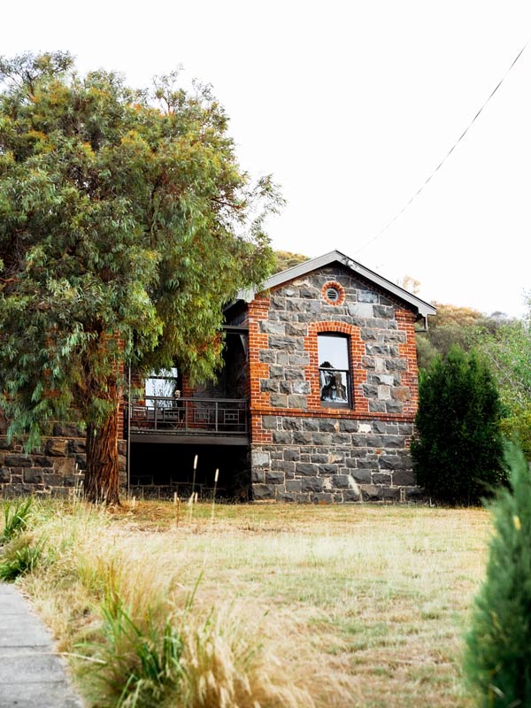 A small cottage with black and red bricks and windows next to a tree on a grass reserve.