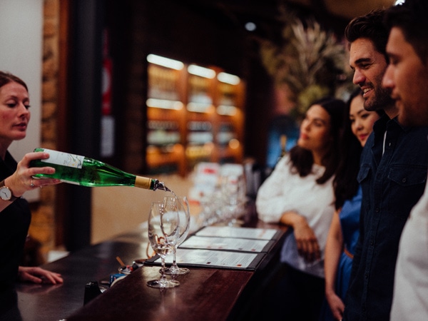 A woman pouring wine from a wine bottle into a glass with two women and two men looking at it.