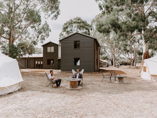 Two men and a baby sitting on chairs in the middle of tents and a wooden house.