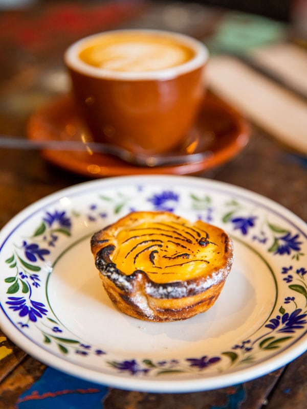 A yellow pastry on a white and blue plate with a coffee in a brown mug in a wooden table.