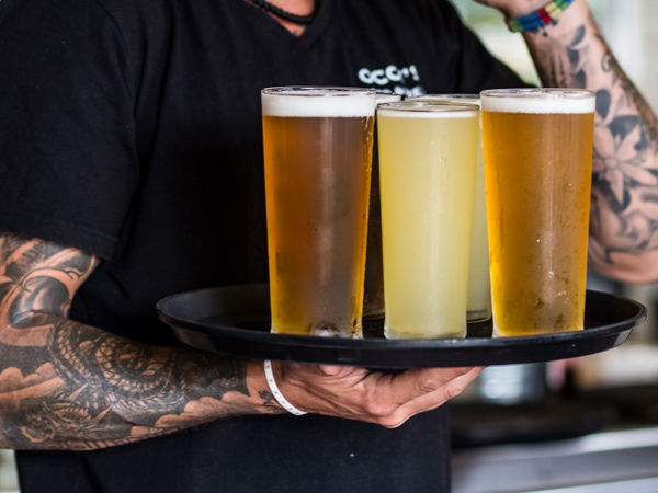 A tattooed man holding a plate with three beers. 