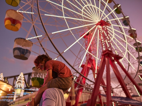 a child plays piggyback on his dad with a Ferris wheel on the background at Luna Park Sydney, Milsons Point