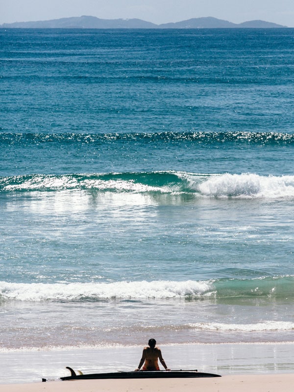 a surfer sitting on a surfboard on the shore while watching the waves