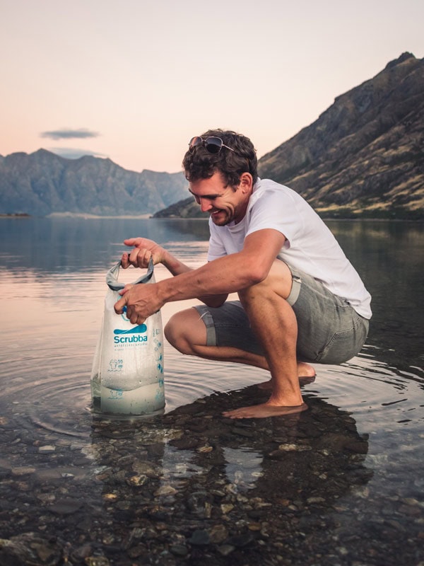 A man using the scrubba kit to clean in a lake by the mountains