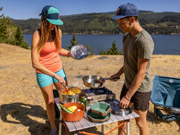 A man and women making a meal on the beach with Gerbers camping gear.