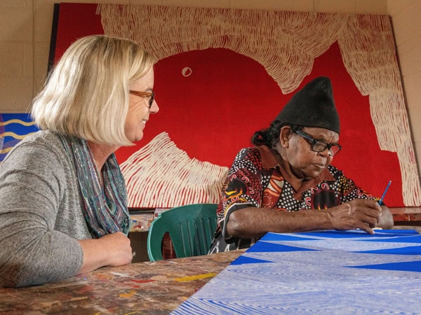 a woman watching an Aboriginal Artist working on a painting at Ikuntji Artists, Haasts Bluff