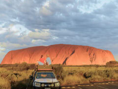 couple sit on top of their car in front of uluru