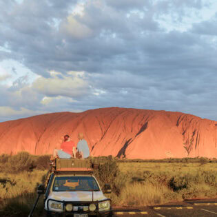 couple sit on top of their car in front of uluru