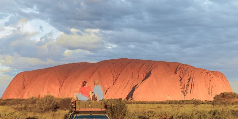 couple sit on top of their car in front of uluru
