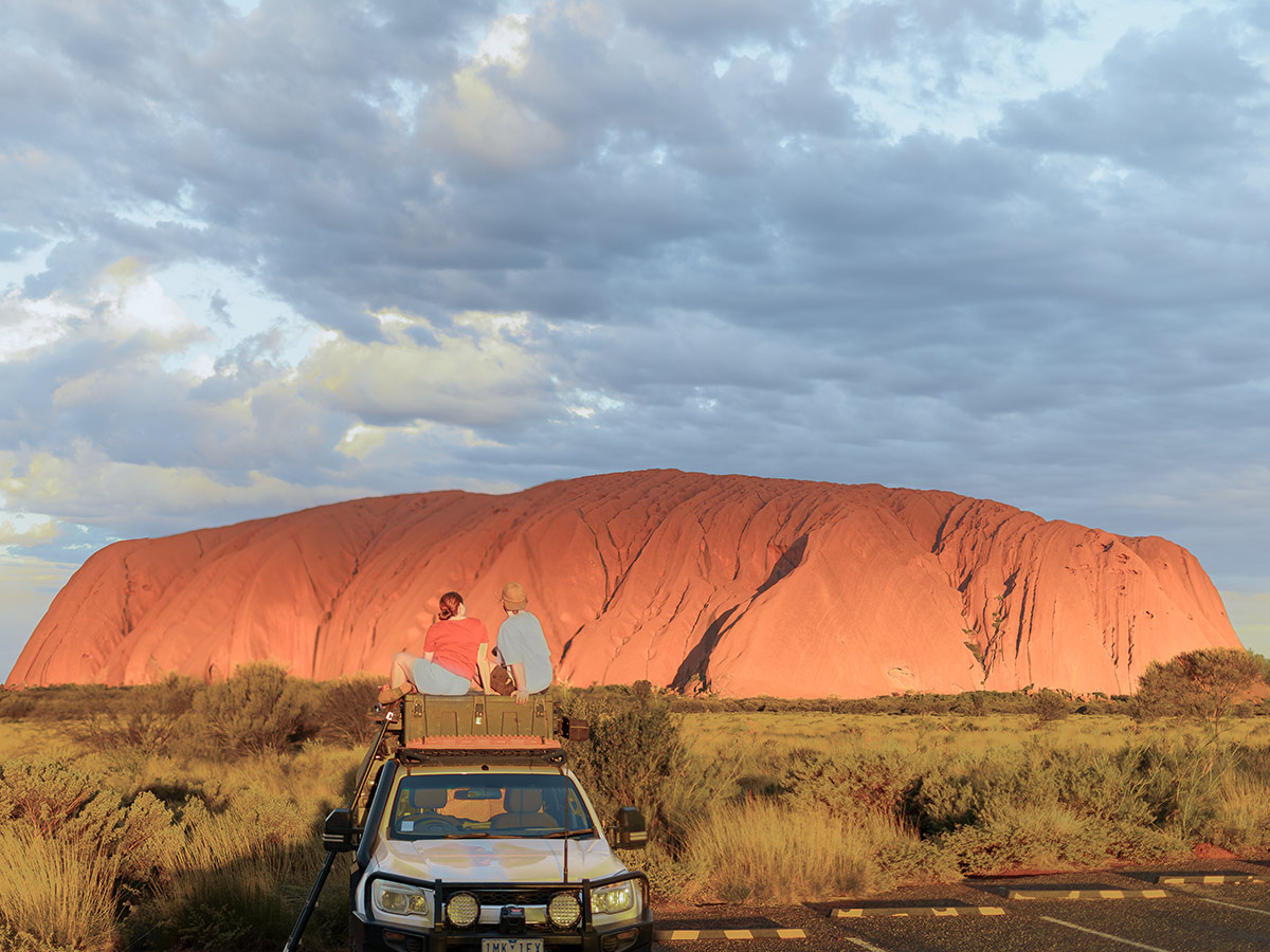 couple sit on top of their car in front of uluru