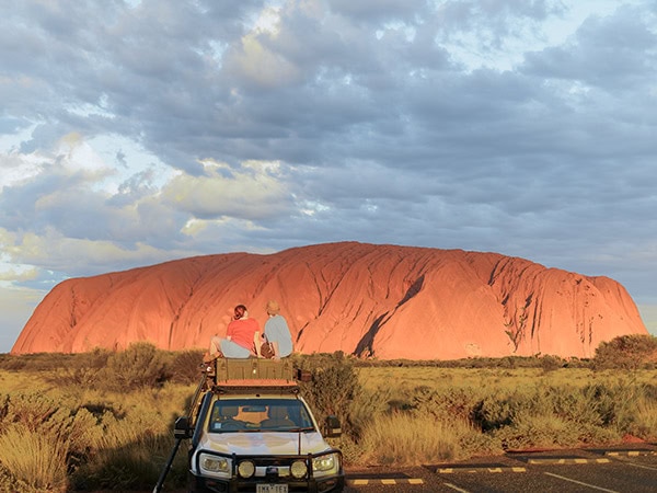 couple sit on top of their car in front of uluru