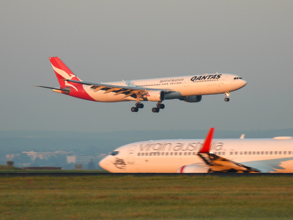 A Qantas Airbus A330-303 plane, registration VH-QPB, coming into land from the south on the main runway of Sydney Kingsford-Smith Airport as flight QF128 from Hong Kong. In the foreground is a Virgin Australia Boeing B737-8FE plane, registration VH-YFZ, taxiing before departure as flight VA1528 to Hobart. 