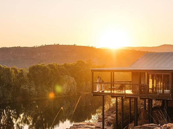 Two people stand beside a suite perched on the edge of a striking burnt-orange cliff, gazing out over the serene Chamberlain River below.