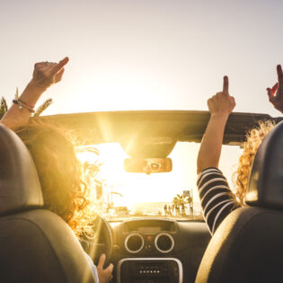Couple of woman friends traveling and driving having a lot of fun dancing in the car with opened roof and summer vacation sunset ocean in front