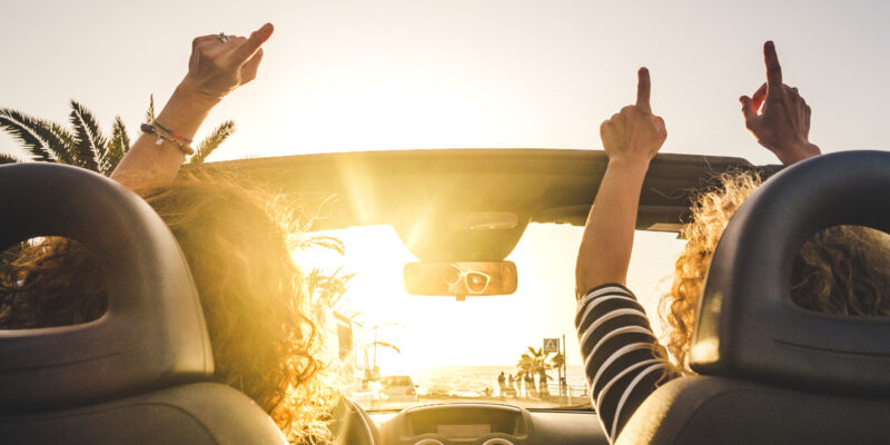 Couple of woman friends traveling and driving having a lot of fun dancing in the car with opened roof and summer vacation sunset ocean in front