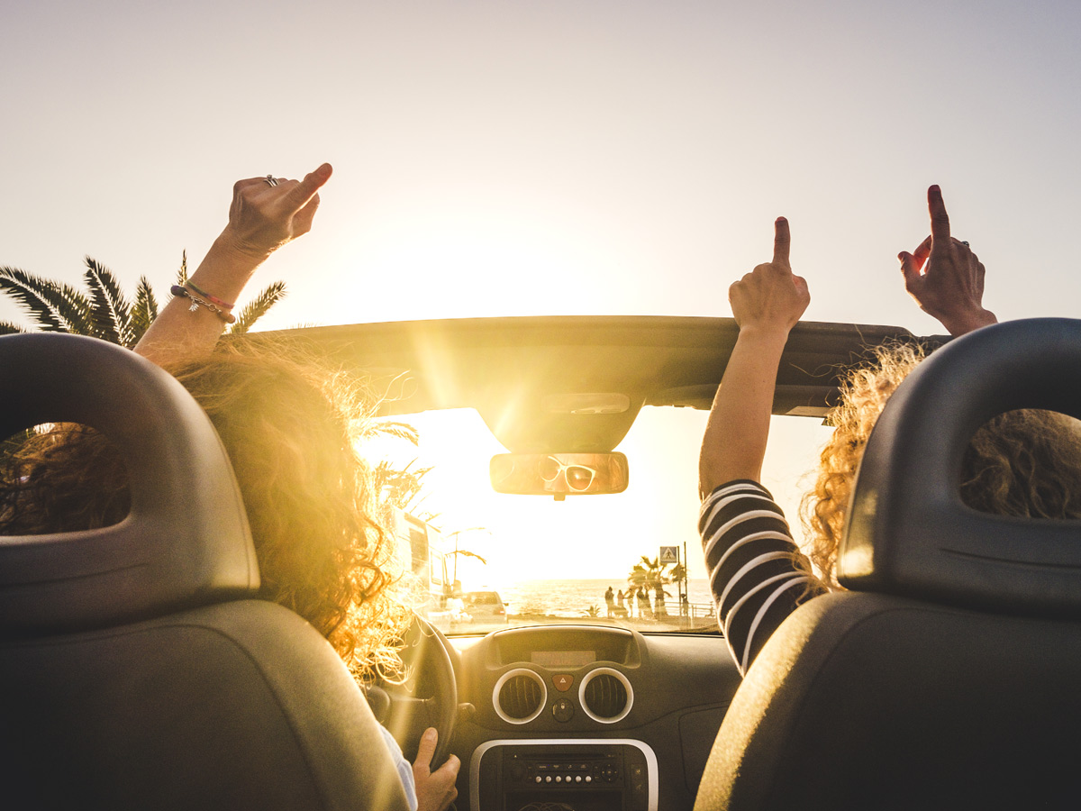 Couple of woman friends traveling and driving having a lot of fun dancing in the car with opened roof and summer vacation sunset ocean in front